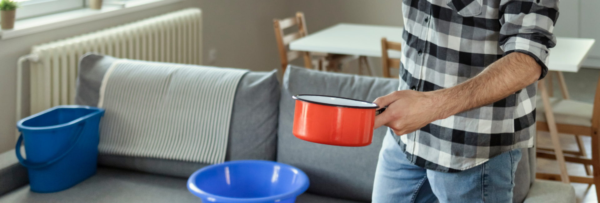 Image of man holding pot in one hand and phone in other hand under a roof leak.